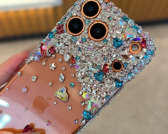 May include: A clear phone case bedazzled with a variety of colorful rhinestones and gems. The case features a gradient design with a concentration of gems around the camera lenses. The gems include clear, blue, red, and iridescent stones.