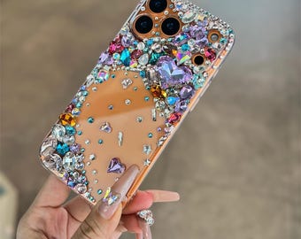 May include: A clear phone case adorned with an array of colorful rhinestones and gems. The embellishments include various shapes and sizes of crystals in shades of blue, purple, green, and clear, creating a sparkling, decorative accessory.