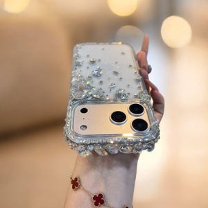 Puede incluir: Funda transparente para teléfono con adornos de cristal plateado y transparente. La funda tiene un panel de cámara plateado con tres lentes de cámara negras y una lente pequeña. La funda se sostiene en una mano y se ve una pulsera roja y dorada.