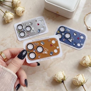 May include: Decorative phone camera lens protectors in silver, brown, and blue. Each protector features a series of circular lens openings and is embellished with sparkling crystals and gems. The text "IP17PRO MAX" is printed on each protector.