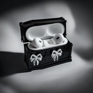 以下が含まれることがあります： 白いリボンがアクセントの黒い棺型のイヤホンケース。ケースは開いており、中に2つの白いイヤホンが見えます。ワイヤレスイヤホンを保護し、保管するためのユニークなアクセサリーです。