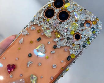 May include: A bedazzled phone case with a peach-colored background. The case is covered in a variety of sparkling rhinestones and crystals in clear, yellow, and iridescent colors. A red heart-shaped gem is visible.