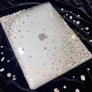 May include: Clear laptop case with a sparkling design. The case is adorned with numerous clear, faceted rhinestones, concentrated along the edges and scattered across the surface. The Apple logo is visible.
