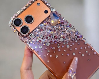 May include: A rose gold phone case adorned with an array of sparkling rhinestones and crystals. The case features a gradient design, transitioning from a dense cluster of gems at the top to scattered accents at the bottom. The camera lenses are outlined with silver.