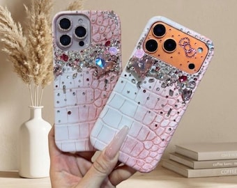 May include: Two bedazzled phone cases. The cases have a pink and white crocodile skin pattern. The top of the cases are decorated with rhinestones and gems. The phone cases are held in a person's hand.
