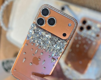 May include: A clear phone case with a peach-colored back, embellished with sparkling rhinestones. The top section features a cluster of large, clear crystals, while smaller gems cascade down the case. The camera lenses are surrounded by a ring of rhinestones.
