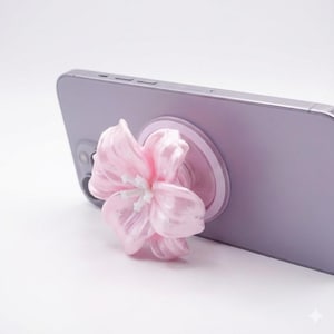 May include: A lilac smartphone with a pink flower-shaped phone grip attached. The flower has a white centre and is made of a translucent material. The phone grip is circular and matches the phone's colour.