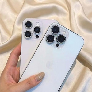 Op de afbeelding: Twee witte smartphones met cameralensbeschermers. De beschermers hebben een zilveren rand en zijn ingelegd met kleine, heldere strass-steentjes. De telefoons worden tegen een achtergrond van een zijdeachtige, crèmekleurige stof gehouden.