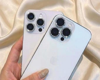 Puede incluir: Dos teléfonos inteligentes blancos con protectores de lente de cámara. Los protectores tienen un borde plateado y están incrustados con pequeños diamantes de imitación transparentes. Los teléfonos se sostienen contra un fondo de tela sedosa de color crema.