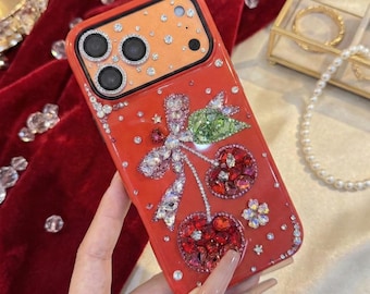 Puede incluir: Una funda de smartphone de color naranja brillante con un motivo decorativo de cerezas hecho de cristales rojos y verdes. La funda está adornada con numerosos diamantes de imitación transparentes y un diseño de lazo incrustado de cristales.