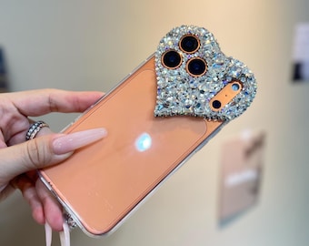 May include: A clear phone case with a peach-colored back. The camera area is embellished with a heart-shaped design encrusted with sparkling rhinestones. The case has a silver-colored trim.