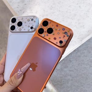 Puede incluir: Dos fundas decorativas para teléfono. Una es plateada con pedrería transparente, la otra es cobriza con pedrería colorida. Ambas fundas tienen un acabado brillante y presentan el logotipo de Apple.