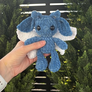 May include: A plush crochet creature made of blue yarn with a soft, fuzzy texture. It features large, dark, round eyes, prominent floppy ears, and white, textured wings extending from its back. The figure has a rounded head and limbs.