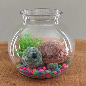 May include: A clear glass terrarium with a plush green and brown turtle, artificial greenery, and colorful gravel. The turtles are inside the glass bowl, which sits on a wooden surface.