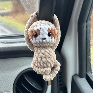 May include: A crocheted sloth hanging from a car's rearview mirror. The sloth is brown and white with black eyes and a brown nose.