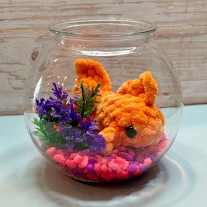 May include: A glass fishbowl with a bright orange crocheted fish, pink and purple pebbles, and a small purple flower arrangement.