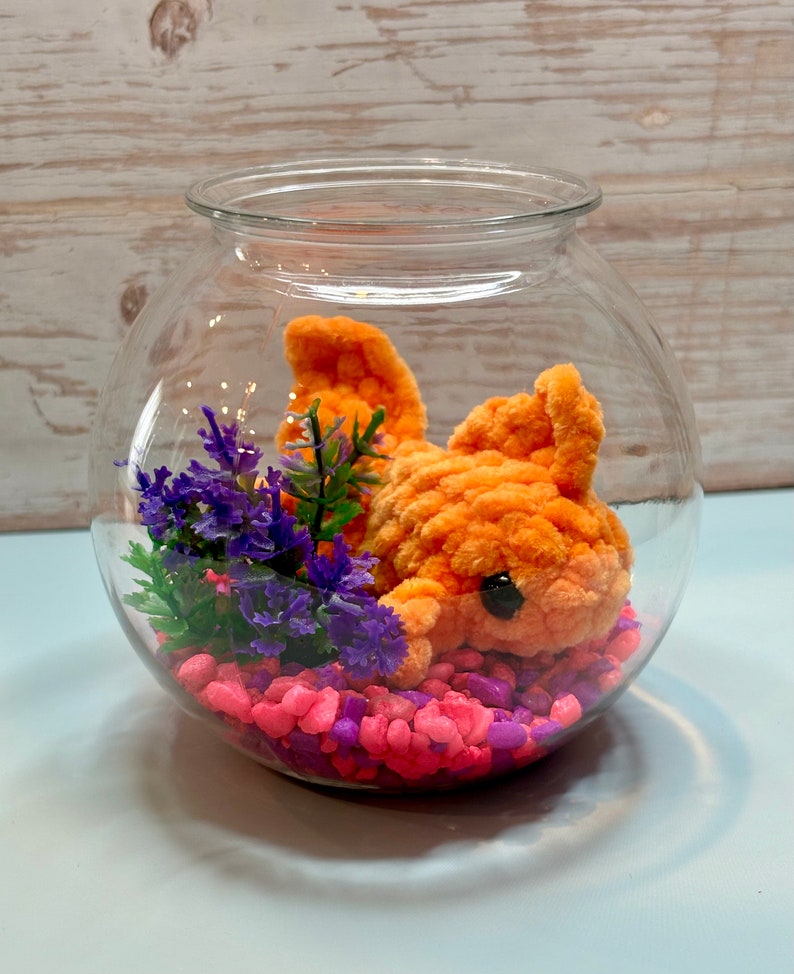 May include: A glass fishbowl with a bright orange crocheted fish, pink and purple pebbles, and a small purple flower arrangement.