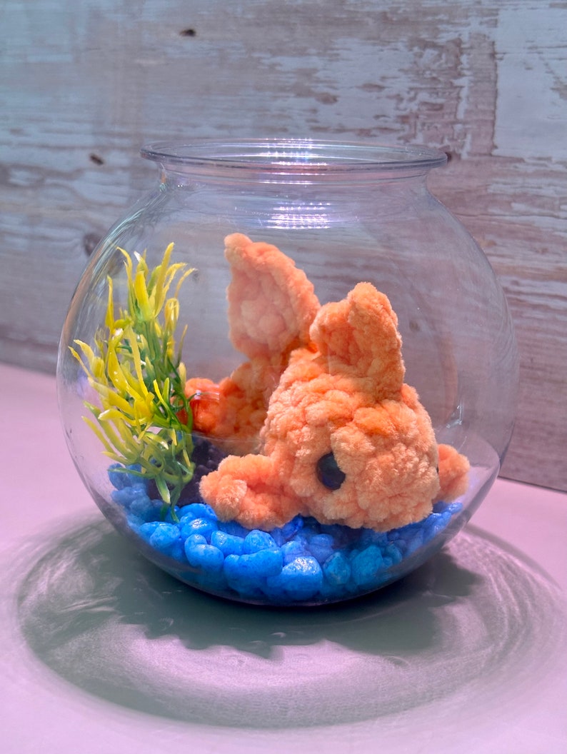 May include: A plush orange fish in a glass bowl with blue pebbles and a yellow plant.