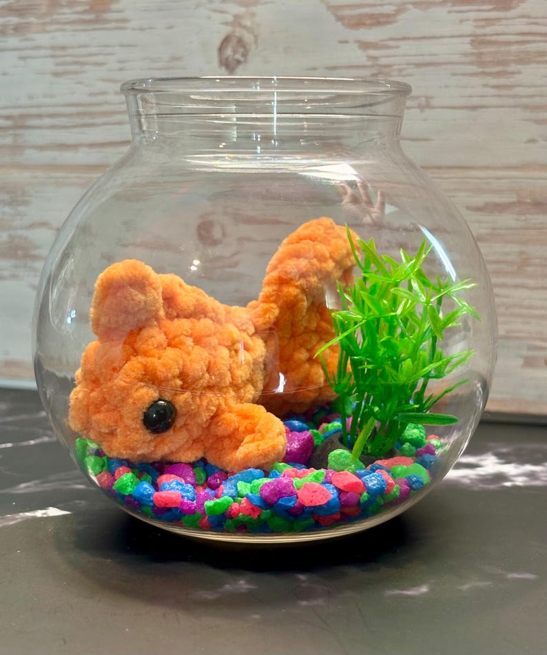 May include: A crocheted orange goldfish in a glass bowl with green plastic plants and colorful pebbles.