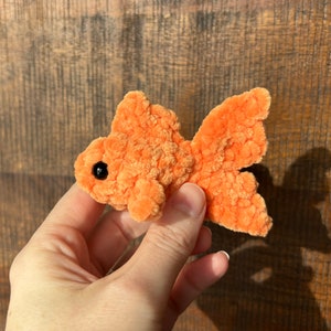 May include: A small, orange, crocheted goldfish with black eyes. The goldfish is being held in a hand.