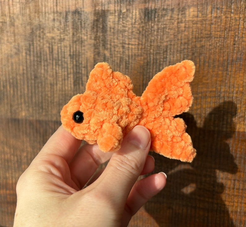 May include: A small, orange, crocheted goldfish with black eyes. The goldfish is being held in a hand.