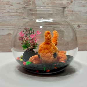 May include: A clear glass globe terrarium with a small orange fuzzy creature, a small green plant with pink flowers, and black and colorful pebbles.
