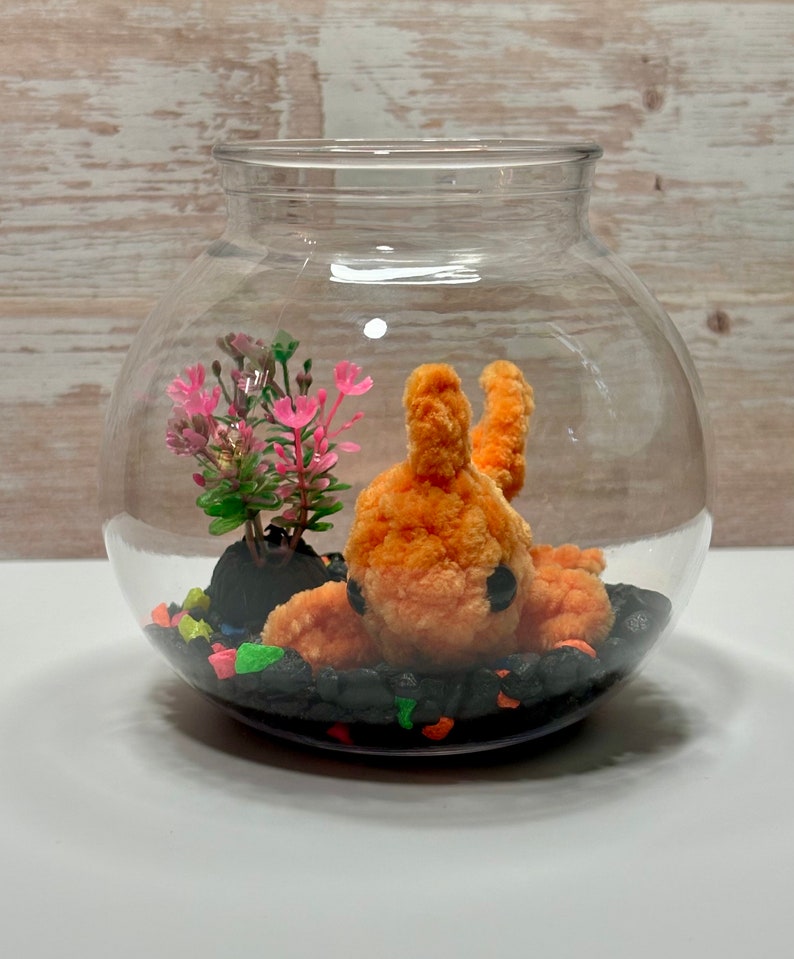May include: A clear glass globe terrarium with a small orange fuzzy creature, a small green plant with pink flowers, and black and colorful pebbles.