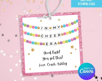 May include: A pink gift tag with a glittery border and a white string. The tag says "IN MY CHEER ERA" with colorful bead garlands. It includes the text "Good luck! You got this! Love, Coach Ashley."