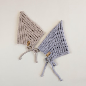 Könnte beinhalten: Zwei gestrickte Baby-Mützen, eine in Beige und eine in Helllila, mit einem kleinen Etikett mit der Aufschrift "Baby" auf jeder.