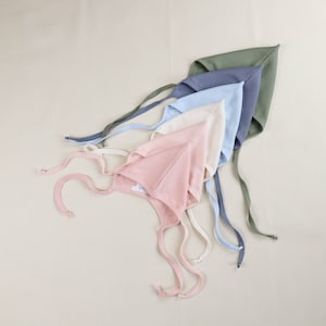Könnte beinhalten: Eine Auswahl an Baby-Häubchen in Pastelltönen: Rosa, Creme, Hellblau, Blau und Grün. Jede Haube hat eine gerippte Textur und lange Bänder zum Binden unter dem Kinn. Die Hauben sind fächerförmig angeordnet.