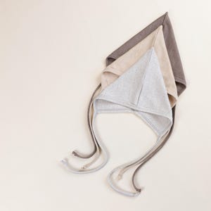 Könnte beinhalten: Drei Baby-Häubchen in neutralen Farben: Hellgrau, Beige und Braun. Jedes Häubchen hat eine dreieckige Form mit langen Bändern zum Binden unter dem Kinn. Die Häubchen sind aus weichem Stoff gefertigt und in einer geschichteten Komposition angeordnet.