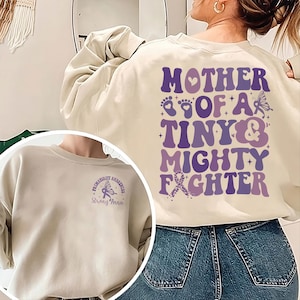 May include: A cream-colored sweatshirt with a purple and pink design that says "Mother of a Tiny & Mighty Fighter" on the back. The front of the sweatshirt has a purple ribbon with a fairy and the words "Prematurity Awareness Strong Anna".