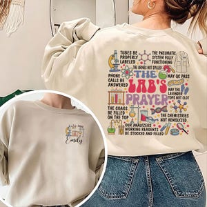 Può includere: Una felpa beige con un design grafico divertente che presenta una preghiera a tema laboratorio. Il testo recita "The Lab's Prayer" e include un elenco di compiti e richieste relative al laboratorio, come "Tubes be properly labeled", "The coag's be filled on the top" e "May the lavender tops not clot". La felpa ha il nome "Emily" stampato sul davanti.