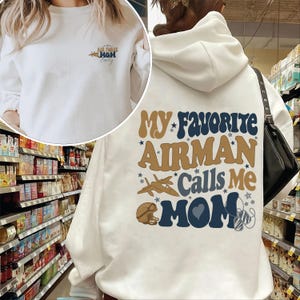 Könnte beinhalten: Weißer Kapuzenpullover mit dem Text "Air Force Mom" auf der Vorderseite und "My Favorite Airman Calls Me Mom" auf der Rückseite. Das Design umfasst ein Flugzeug, Sterne und einen Helm in Blau und Braun.
