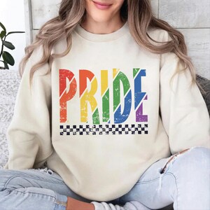 May include: A cream colored sweatshirt with the word "PRIDE" printed in rainbow colors with a checkered pattern underneath.