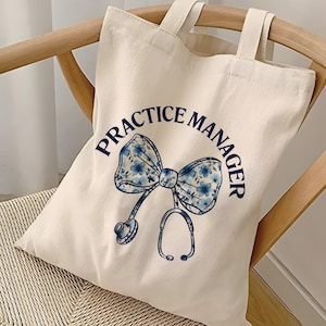 May include: Cream-colored tote bag with the words "PRACTICE MANAGER" in navy blue. A navy blue bow with a floral pattern and a stethoscope design are centered on the bag. The bag has two handles and is sitting on a wooden chair.