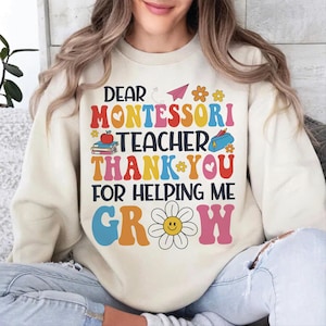 Op de afbeelding: Crèmekleurige sweatshirt met een kleurrijke afbeelding met de tekst "DEAR MONTESSORI TEACHER THANK YOU FOR HELPING ME GROW". Het ontwerp bevat een papieren vliegtuig, bloemen, een appel, boeken en een etui.
