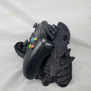 Op de afbeelding: Een zwarte 3D-geprinte drakenkop-controllerstandaard. De standaard is ontworpen om een ​​videogamecontroller vast te houden. De draak heeft zijn bek open en de controller ligt erin.