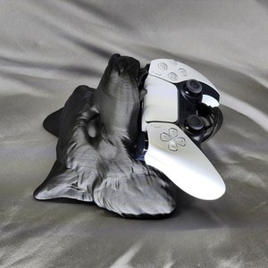 Może przedstawiać: Stojak na kontroler PlayStation 5 w kształcie głowy wilka, wydrukowany w 3D. Stojak jest wykonany z czarnego plastiku i ma realistyczny design głowy wilka.