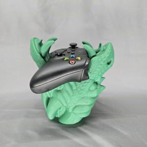 May include: A black Xbox controller rests in a light green, stylized holder. The holder has a textured, organic design, possibly resembling a creature's jaws or claws. The controller's buttons are visible.