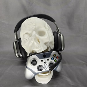 May include: A white skull wearing black and silver headphones and holding a silver and black video game controller. The controller has colored buttons and a headset microphone.