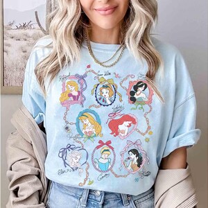 Puede incluir: Camiseta azul claro con retratos de princesas de Disney en marcos ornamentados. La camiseta incluye retratos de Rapunzel, Blancanieves, Jasmine, Ariel, Cenicienta, Elsa y otras. La camiseta es de color azul claro.