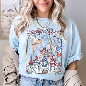 Puede incluir: Camiseta azul claro con una colorida ilustraci&oacute;n de un castillo, fuegos artificiales y Campanilla. El dise&ntilde;o incluye un lazo rojo, blanco y azul, globos de Mickey Mouse y detalles florales. Las mangas est&aacute;n dobladas.