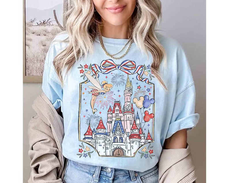 Puede incluir: Camiseta azul claro con una colorida ilustraci&oacute;n de un castillo, fuegos artificiales y Campanilla. El dise&ntilde;o incluye un lazo rojo, blanco y azul, globos de Mickey Mouse y detalles florales. Las mangas est&aacute;n dobladas.