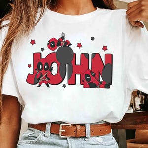 May include: White t-shirt with the name "JOHN" in red block letters, decorated with Deadpool illustrations. The design includes Deadpool characters, stars, and a black wrecking ball. The shirt has a brown belt.