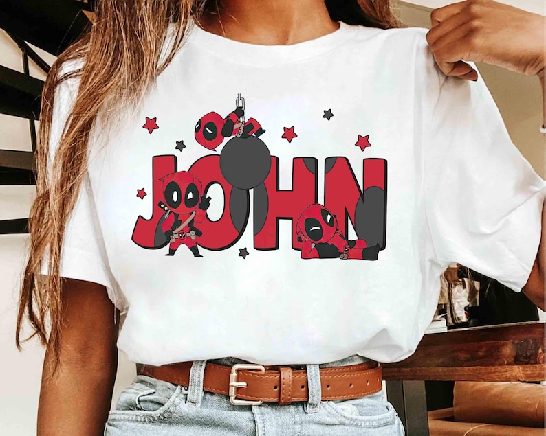 May include: White t-shirt with the name "JOHN" in red block letters, decorated with Deadpool illustrations. The design includes Deadpool characters, stars, and a black wrecking ball. The shirt has a brown belt.