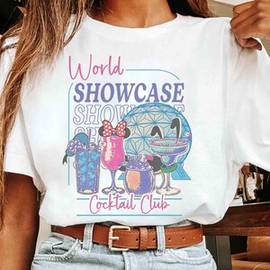 以下が含まれることがあります： グラフィックデザインが施された白いTシャツ。「World Showcase Cocktail Club」の文字とカクテルのイラスト、幾何学的な球体が描かれています。Tシャツは人が着ています。