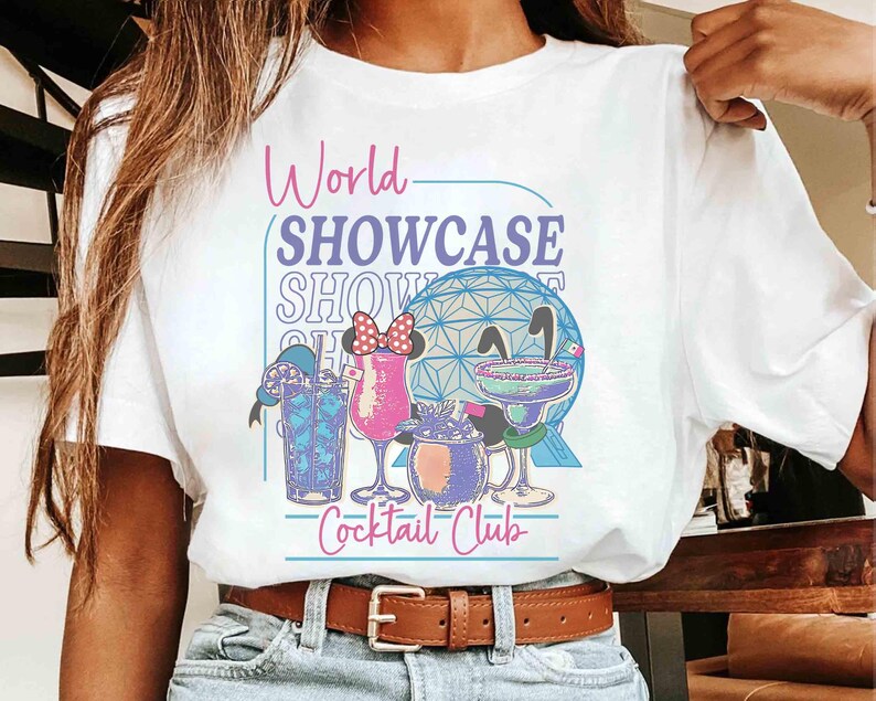 以下が含まれることがあります： グラフィックデザインが施された白いTシャツ。「World Showcase Cocktail Club」の文字とカクテルのイラスト、幾何学的な球体が描かれています。Tシャツは人が着ています。
