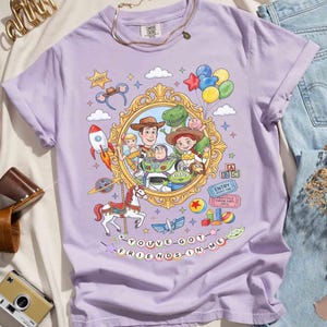 May include: A light purple t-shirt featuring a colorful graphic of characters from a popular animated movie. The design includes a gold-framed portrait, a rocket, a carousel horse, and the text "YOU'VE GOT FRIENDS IN ME."
