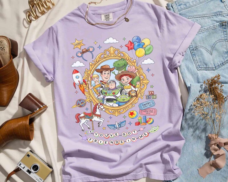 May include: A light purple t-shirt featuring a colorful graphic of characters from a popular animated movie. The design includes a gold-framed portrait, a rocket, a carousel horse, and the text "YOU'VE GOT FRIENDS IN ME."